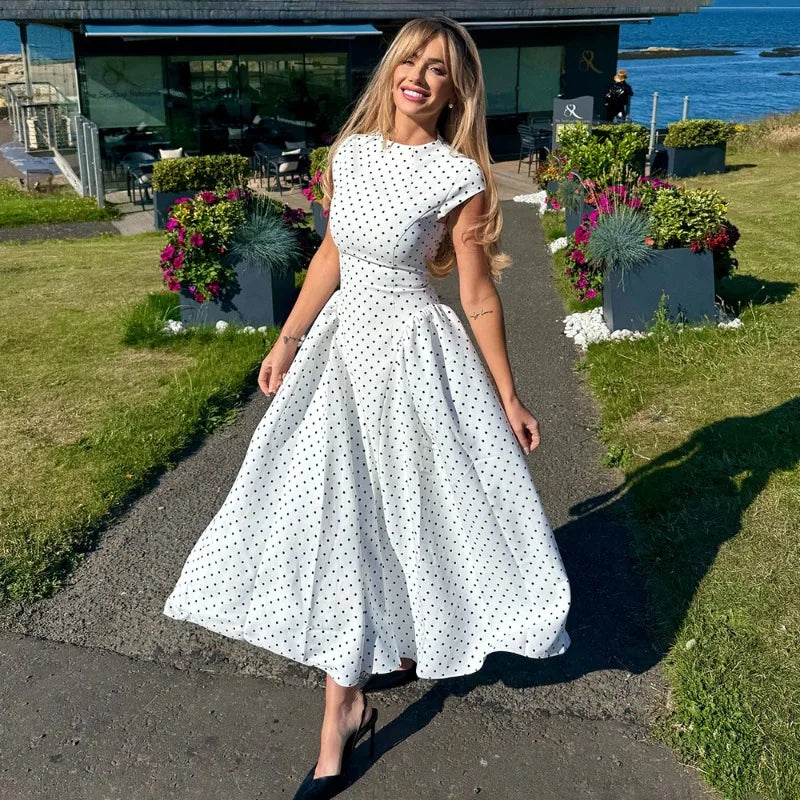 Woman in a white polka dot dress standing on a path with flowers and a building in the background.